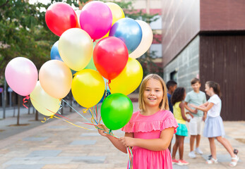 Fototapeta na wymiar Portrait of smiling cute blonde preteen girl standing on city street on summer day, holding bunch of colorful balloons