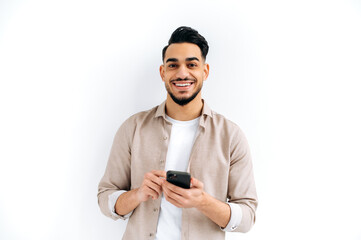 Handsome positive Arabian or Indian modern guy, holding smartphone in his hands, messaging, looking...