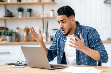 Confused puzzled indian or arabian guy, freelancer or student, sits at home in the kitchen, looking disappointment at the laptop screen, stressed from the failure of a deal or project, in desperation