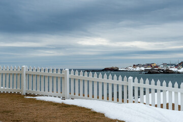 A white wooden picket fence divides and identifies a property boundary corner. There's grass on the ground and white snow. The blue ocean is in the background with a rocky coastline under cloudy skies