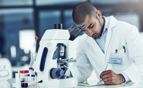 Focused Male Lab Technician Using Digital Tablet Working On Medical Development And Innovation Project With Microscope, Taking Notes. Doctor Diagnosing Test Tube Blood Sample With Science Equipment.