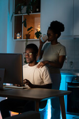 Obraz na płótnie Canvas Young African American couple stays up late due to work on new profitable project. Woman drinks coffee putting hand on shoulder of focused man in glasses sitting at kitchen table near computer closeup