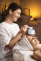 Smiling beautician applying cream to a beautiful woman's face in the spa salon. Health care, beauty...