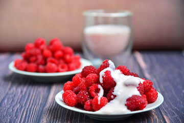 Berries still-life. Heap of red ripe juicy fresh sweet raspberries filled with milk cream and glass cup of yogurt on wooden boards on blurred background. Selective focus