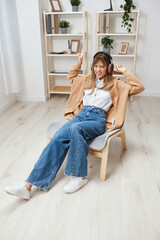 Fototapeta premium Overjoyed cute blonde student lady in warm sweater in headphones listen fav songs flirting at camera sitting in armchair at modern home interior. Music time Relaxing Cool playlist Concept. Copy space