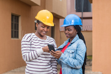 African ladies, women or coworkers from Nigeria with safety helmet, holding and using a phone as...
