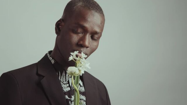 Studio portrait of handsome young adult Black man wearing elegant outfit posing on camera with bunch of fresh flowers enjoying their smell
