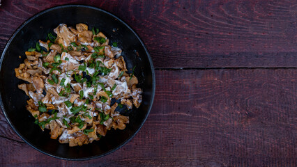 French-fried chanterelle mushrooms with onions and sour cream in a frying pan on a dark wooden background, garlic, onion and parsley and dill greens are laid out next to each other.
