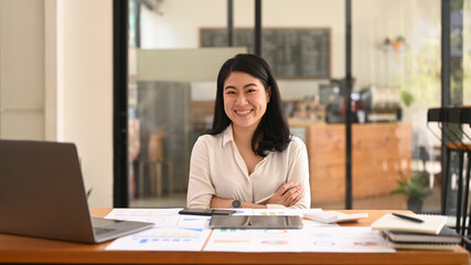 Confident asian woman employee sitting with arms crossed at her workplace and smiling to camera