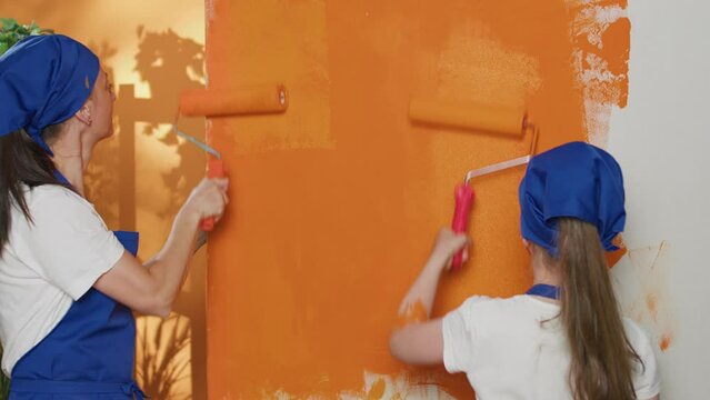 Small family painting apartment walls with orange color, doing renovation work to redecorate house room. Mother and girl using paint and diy tools to renovate home interior. Handheld shot.