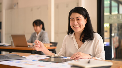 Happy asian female office worker sitting in corporate office ad smiling to camera