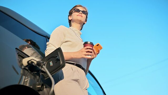 A young blonde woman with smartphone and coffee at a car charging station with charging electric car nearby in Chisinau, Moldova