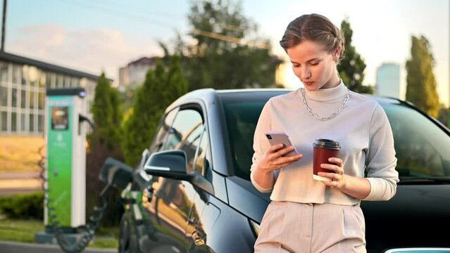 A young blonde woman with smartphone and coffee at a car charging station with charging electric car nearby in Chisinau, Moldova