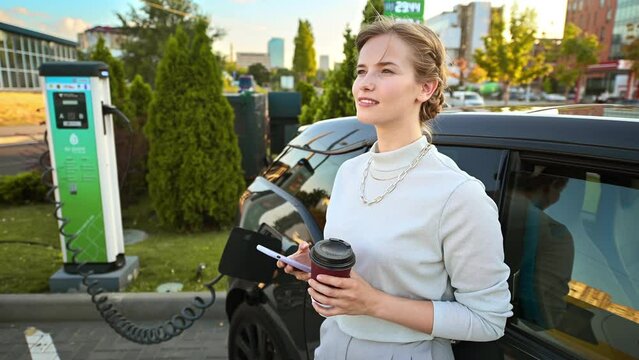 A young blonde woman with smartphone and coffee at a car charging station with charging electric car nearby in Chisinau, Moldova