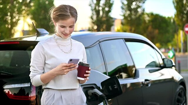 A young blonde woman with coffee using smartphone at a car charging station with electric car nearby in Chisinau at sunset, Moldova. Slow motion