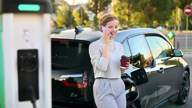 A young blonde woman with coffee talking on the smartphone at a car charging station with electric car nearby in Chisinau at sunset, Moldova