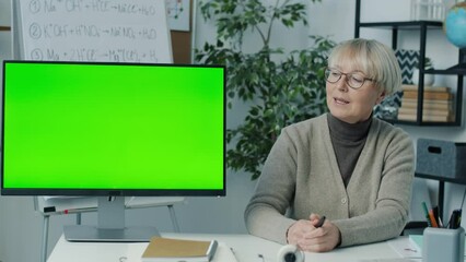Mature lady teaching online courses pointing at computer with chroma key green screen speaking in class. Modern devices and education concept.