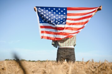 Young patriotic farmer stands among new harvest. Boy walking with the american flag on the wheat field celebrating national independence day. 4th of July concept.