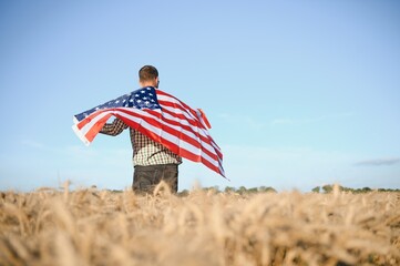 Young patriotic farmer stands among new harvest. Boy walking with the american flag on the wheat field celebrating national independence day. 4th of July concept.