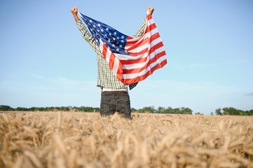 Young patriotic farmer stands among new harvest. Boy walking with the american flag on the wheat field celebrating national independence day. 4th of July concept.