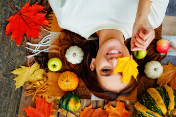 Close-up portrait of a young brunette beautiful woman lying on her back with a closed eye maple leaf. apples, pumpkins and yellow leaves on the hair in the autumn outdoor park. Top view
