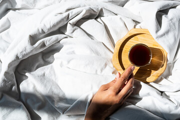 Females hand reaches for morning coffee cup in bed. Woman wakes up on white bedding in sunlight.
