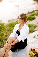 Young attractive girl with dark hair in a white shirt on a picnic in nature by the river in summer at sunset. Healthy life.