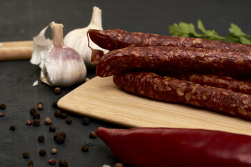 spicy smoked sausages on a cutting board next to red pepper garlic and herbs on a black background