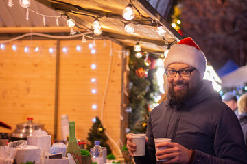 Happy man in a Santa Claus hat and warm clothes at christmas market with hot drinks stall in evening. Christmas winter holidays. Shopping at Christmas Markets