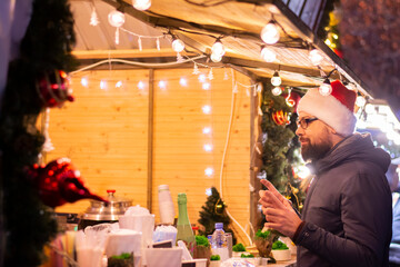 Man has fun at the Christmas market buys drinks and food on the street of the city in the evening of the garland. A man in a warm jacket and Santa Claus hat. Hot winter drinks: mulled wine, cappuccino