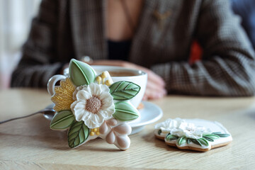 White cup of coffee on saucer and nice icing flower shaped cookies on table on foreground closeup,...