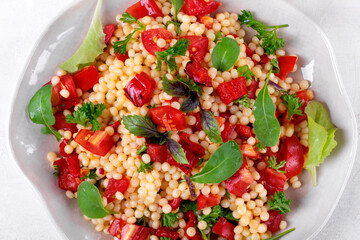 Warm salad with ptitim pasta, grilled peppers, tomato, arugula, basil and olive oil served on the white plate. Top view
