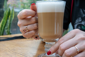 woman hand with beautiful manicure and rings jewelry is holding a cup of coffee latte on a wooden table in the cozy cafeteria. Lifestyle