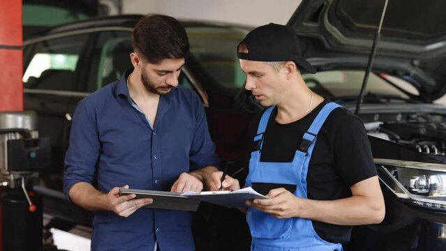 Car service manager with a tablet, talking to mechanic man discussing car diagnostics and repairing. Mechanic talking to manager near vehicle in car service. Specialist is showing info on tablet.