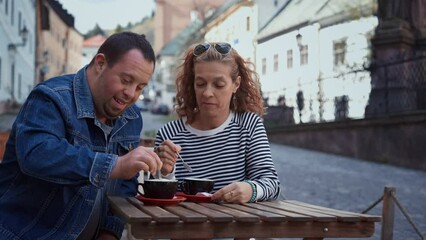 Happy young man with Down syndrome with his mother sitting at cafe outdoors and talking.