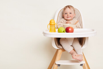 Indoor shot of beautiful cute baby girl wearing beige sweater sitting in a child's chair having dinner or breakfast, , isolated on a white background, copy space for advertisement or promotional text.