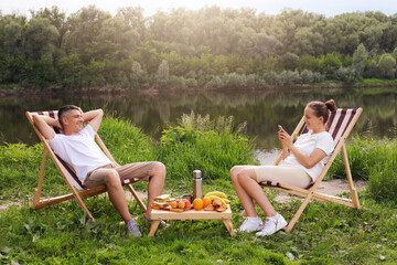 Outdoor shot of young couple sitting on deck chair and talking near the river, relaxed man sits...
