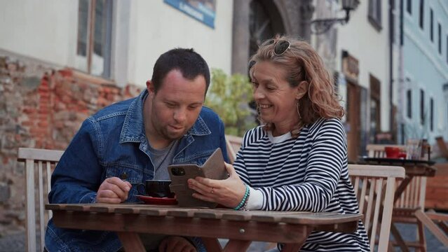 Happy young man with Down syndrome with his mother sitting at cafe outdoors and talking.