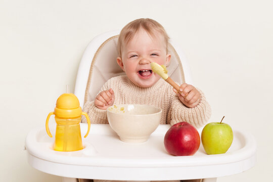 Image of happy positive baby wearing beige sweater sitting in a child's chair eating puree and laughing, holding spoon, posing alone isolated on a white background.