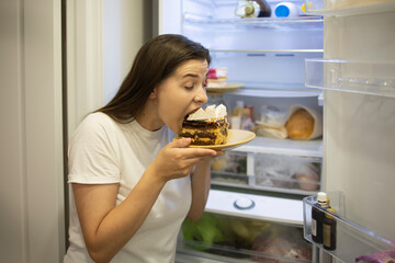 Woman bites a delicious cake at night. Woman in the kitchen has a snack late at night, takes out a...