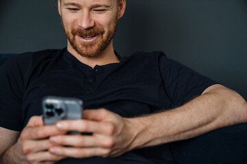 Young ginger man using mobile phone while resting on sofa at home