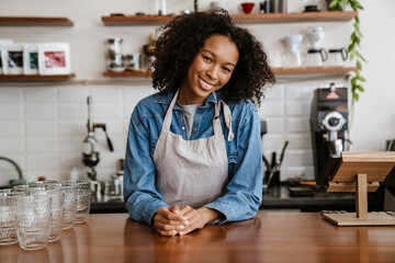 Black young barista woman wearing apron smiling while working in cafe
