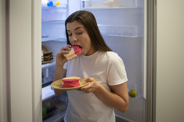 Woman in the sleepwear eating sweet donuts near the refrigerator. Concept: breaking the diet night eating high calorie. Stop diet and gain extra pounds due to carbs food and unhealthy eating