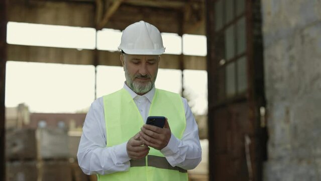 Senior male architect standing outside of building during construction process and typing message on modern smartphone. Positive man in reflective jacket and hardhat chatting online.