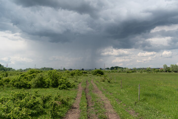 Fototapeta na wymiar Dramatic storm cloud with rain over field with dirt road, stormy weather over pasture