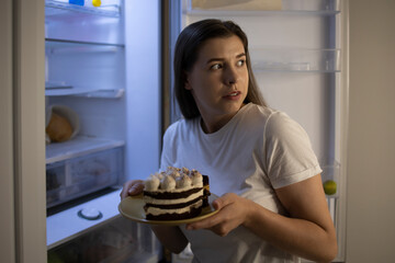 Woman at night in the kitchen the refrigerator with pieces of cake looks around so that no one sees...