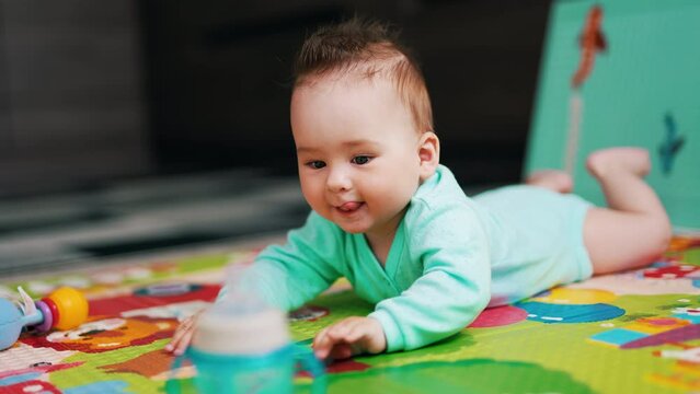 Beautiful baby boy lying on the colorful mat clapping hands by the floor. Funny kid stuck out his tongue and looks at the bottle in front of him.