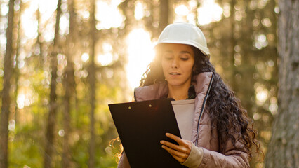 Millennial female technician ecologist looking up at treetops, Young indian woman in hardhat with clipboard taking measures checking trees. Forestry engineer in park. Supervising wildlife sanctuary - obrazy, fototapety, plakaty