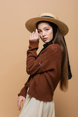 Brunette woman in straw hat and knitted cardigan looking at camera isolated on beige