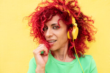 portrait of a latin woman wearing a green t-shirt looking at the camera playing with her red afro hair and listening to music with yellow headphones isolated yellow background
